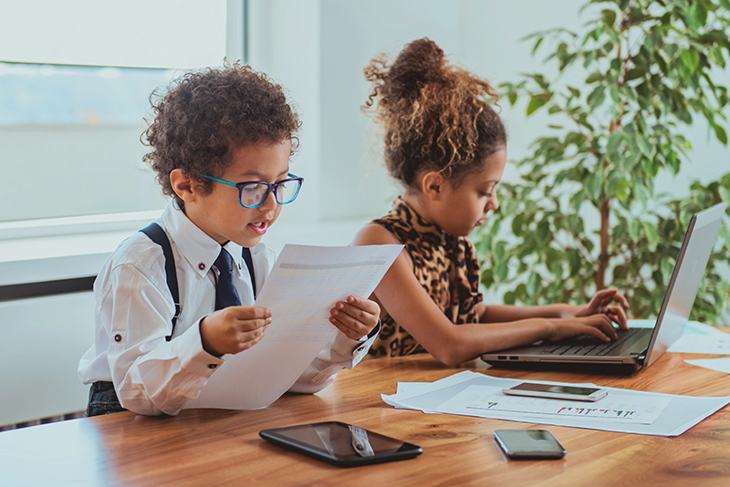 Two children dressed as office workers reading finance documents and working on the computer