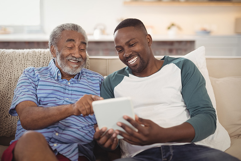 Father sitting on a couch next to adult son looking at a tablet