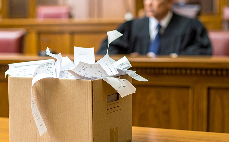Box of unorganized documents in a court room with a judge in the background