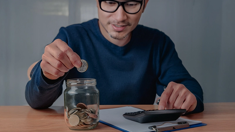 Man dropping money in savings jar with notepad and calculator on his desk